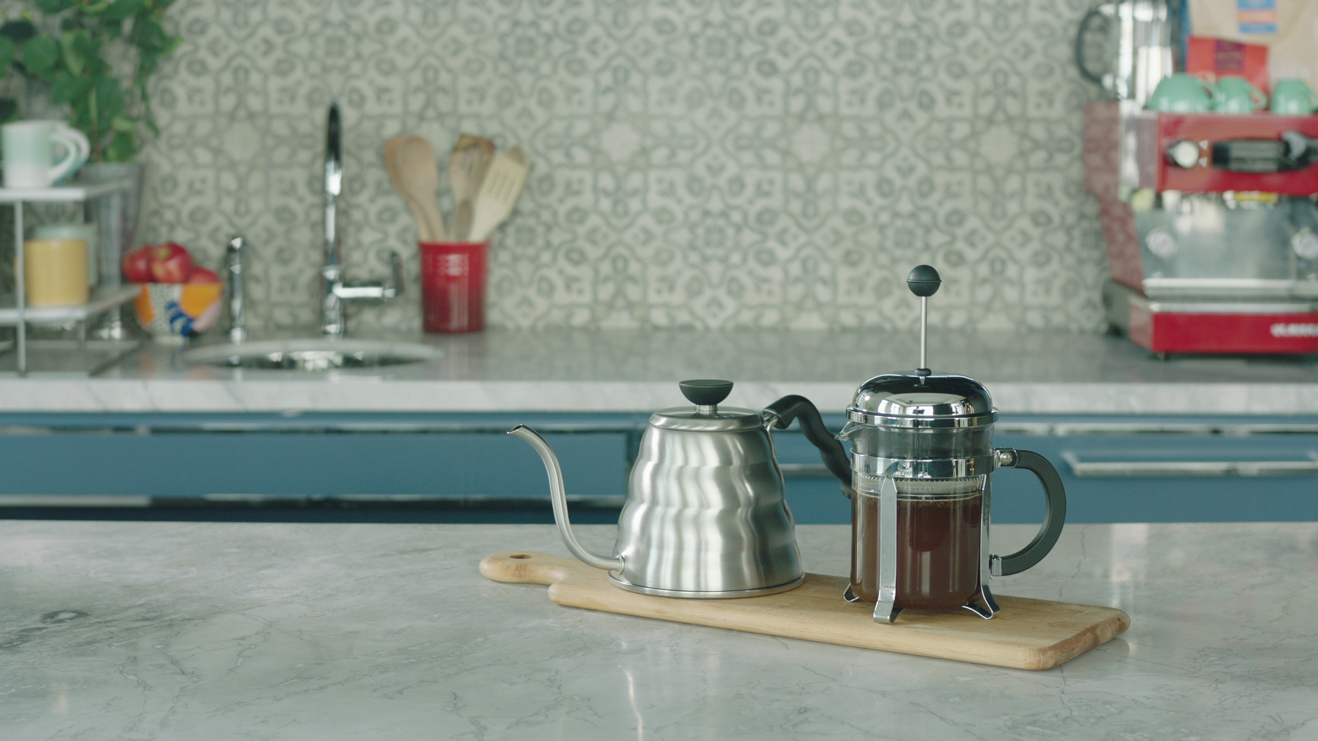 French press brewing coffee beside a stainless steel gooseneck kettle on a cutting board on a marble countertop in a bright kitchen