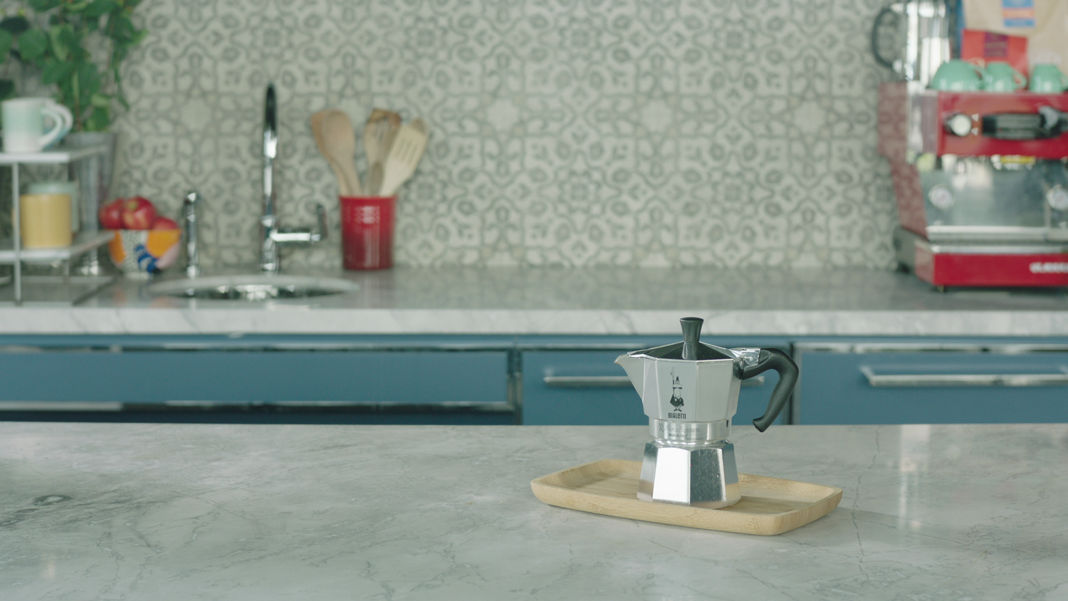 Front view of moka pot showcasing octagonal aluminum body and black handle on a wooden tray on a marble countertop in a kitchen