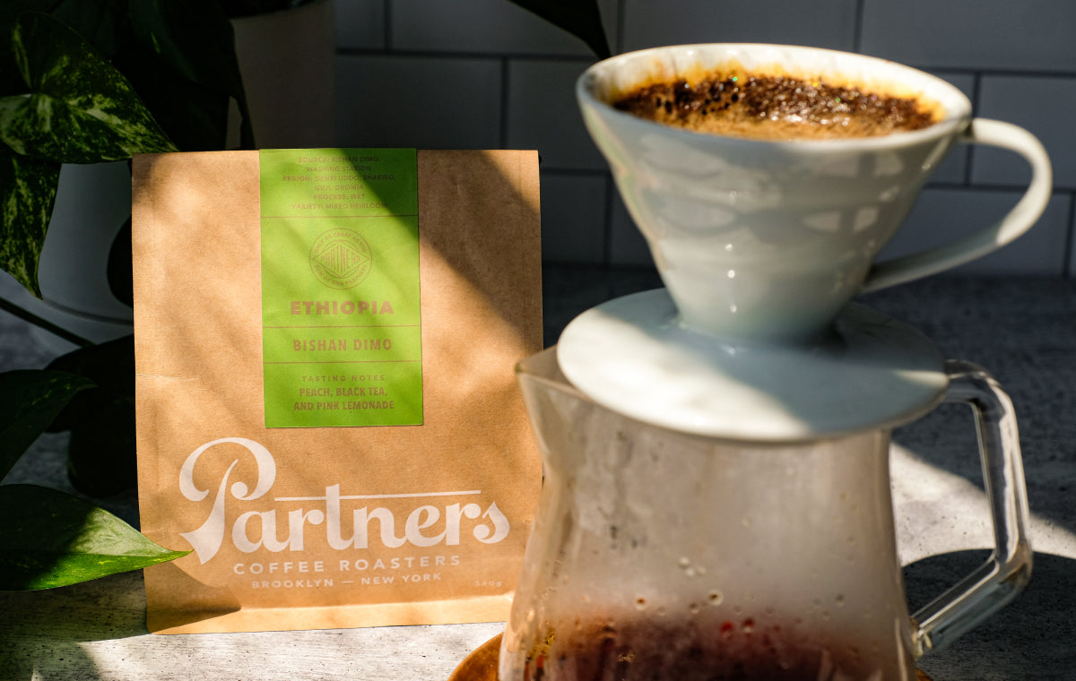 Front of Partners Coffee Ethiopia Bishan Dimo bag with green label beside a ceramic pour-over and carafe on a sunlit counter