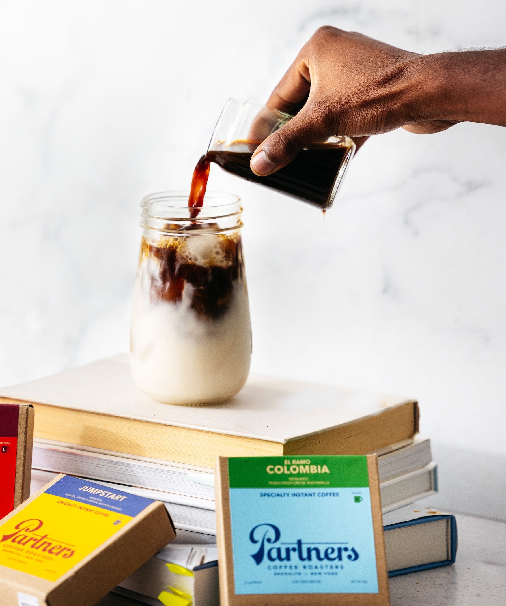 Hand pouring coffee into a milk-filled glass jar over stacked books to make an iced latte