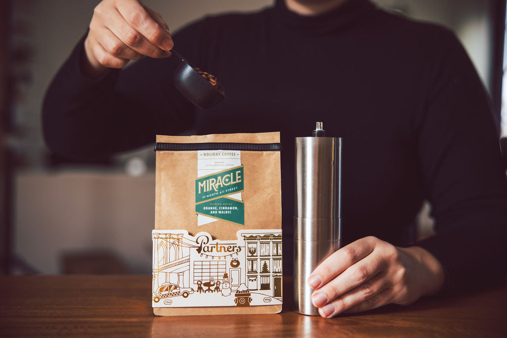 Front view of Miracle holiday coffee bag with Partners storefront illustration, hand scooping beans and stainless grinder