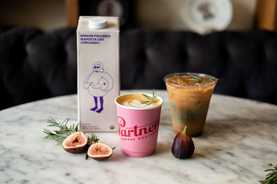 Front view of Partners Coffee hot and iced lattes with Minor Figures Barista Oat carton, figs and rosemary on a marble table