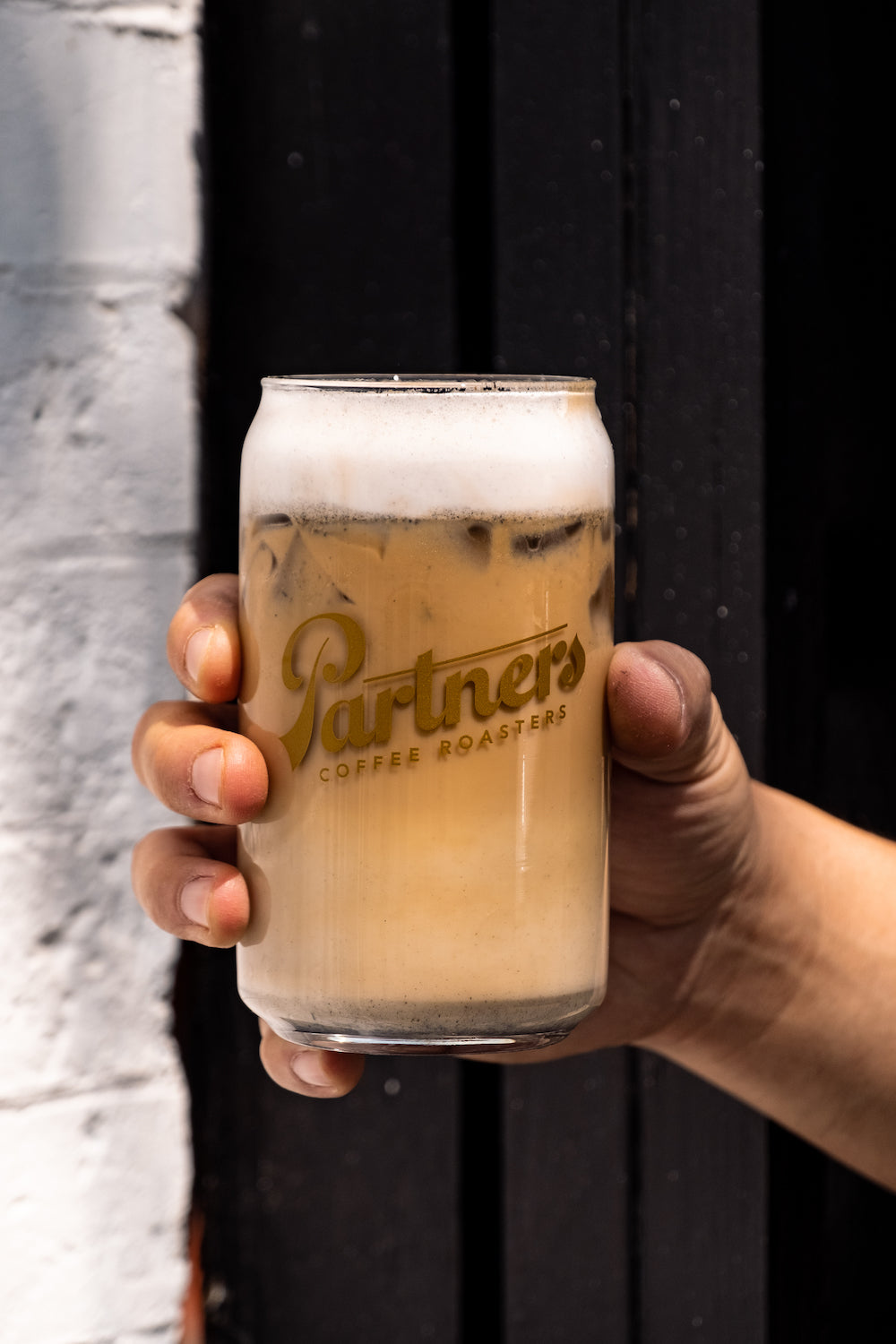Front view of black sesame cold brew latte in Partners glass with creamy foam and sesame sediment, held against dark wood
