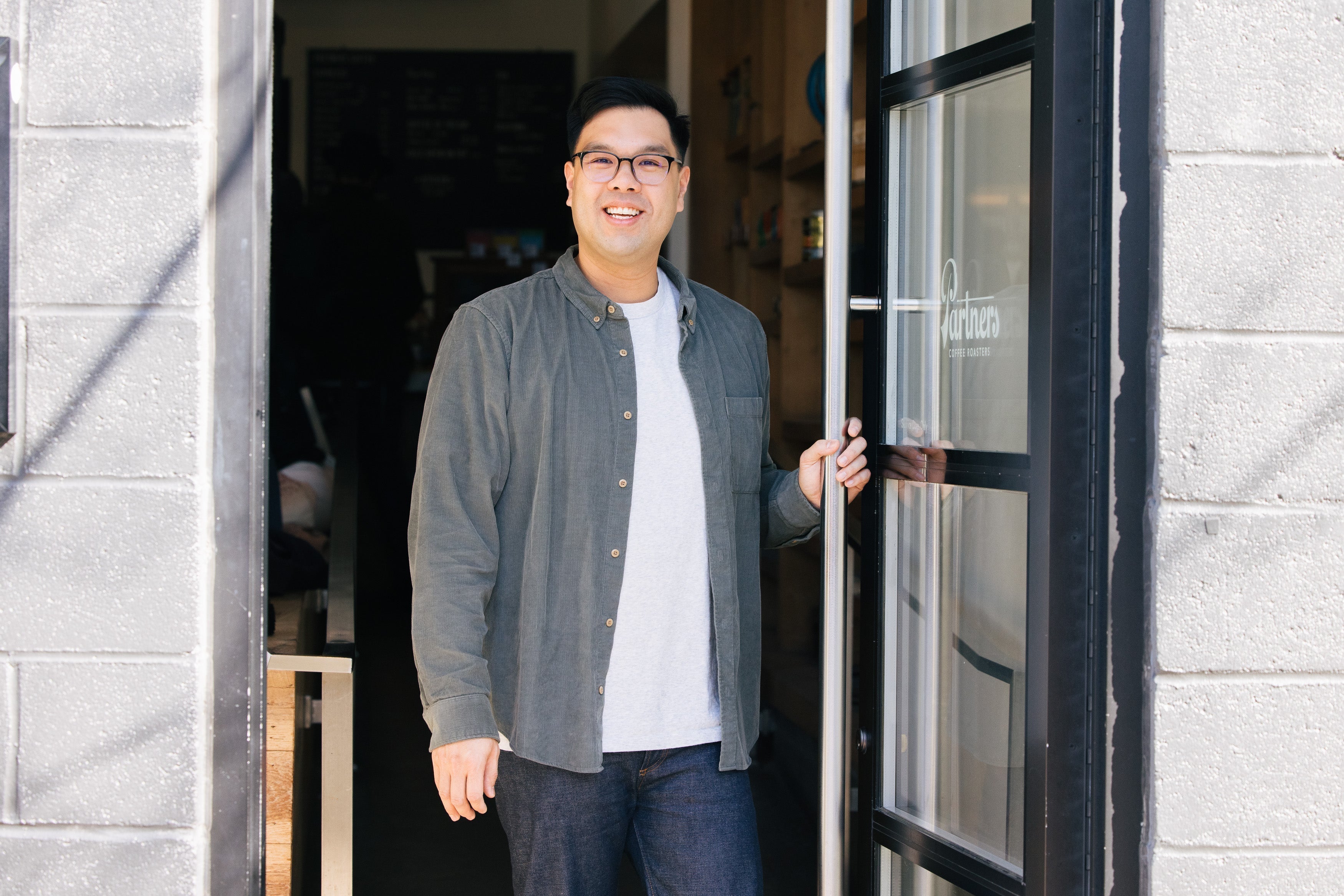 Cary W., Partners Coffee Director of Coffee, smiling as he holds open the cafe’s glass storefront door