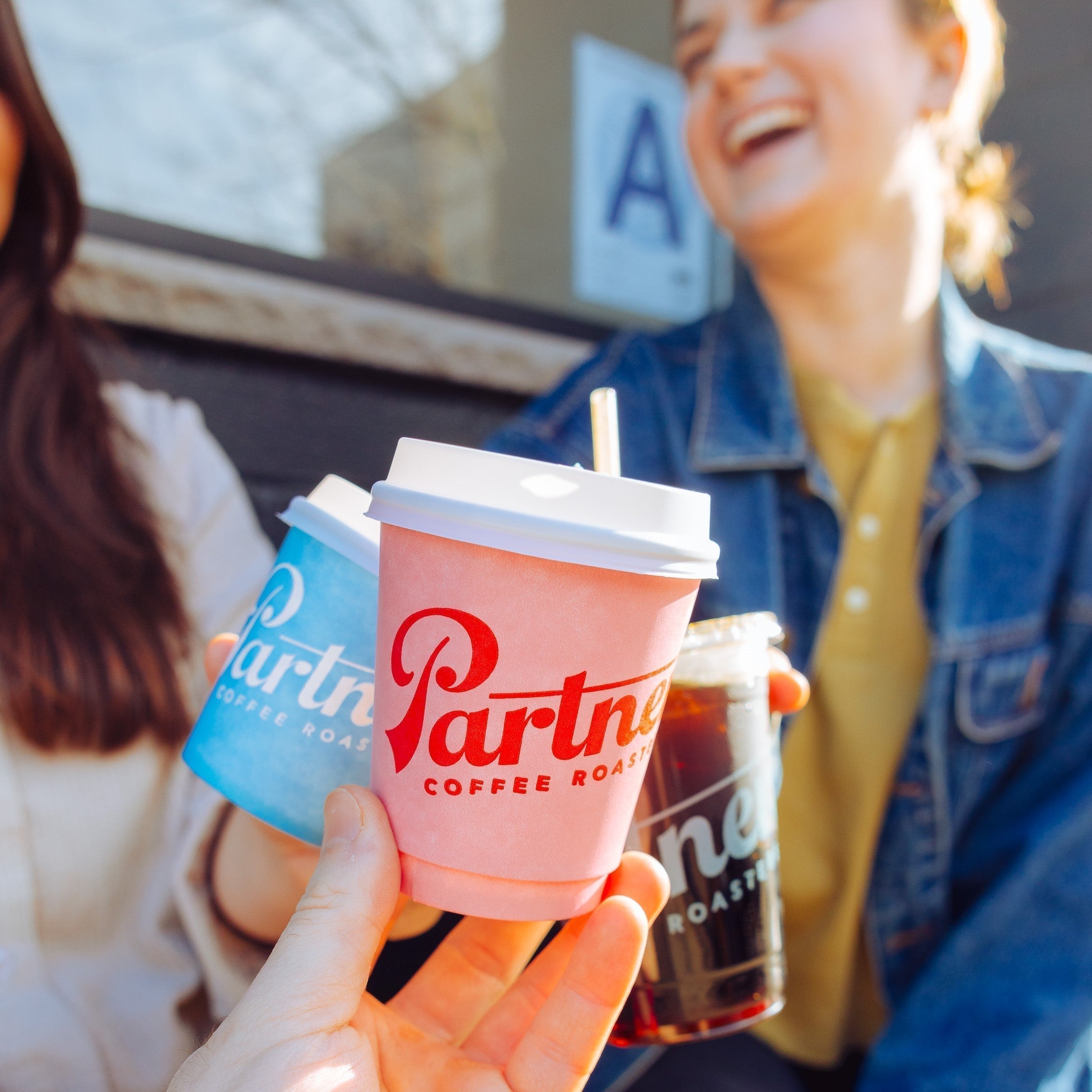 Three friends holding Partners Coffee cups and laughing outside a Partners Coffee shop on a sunny sidewalk