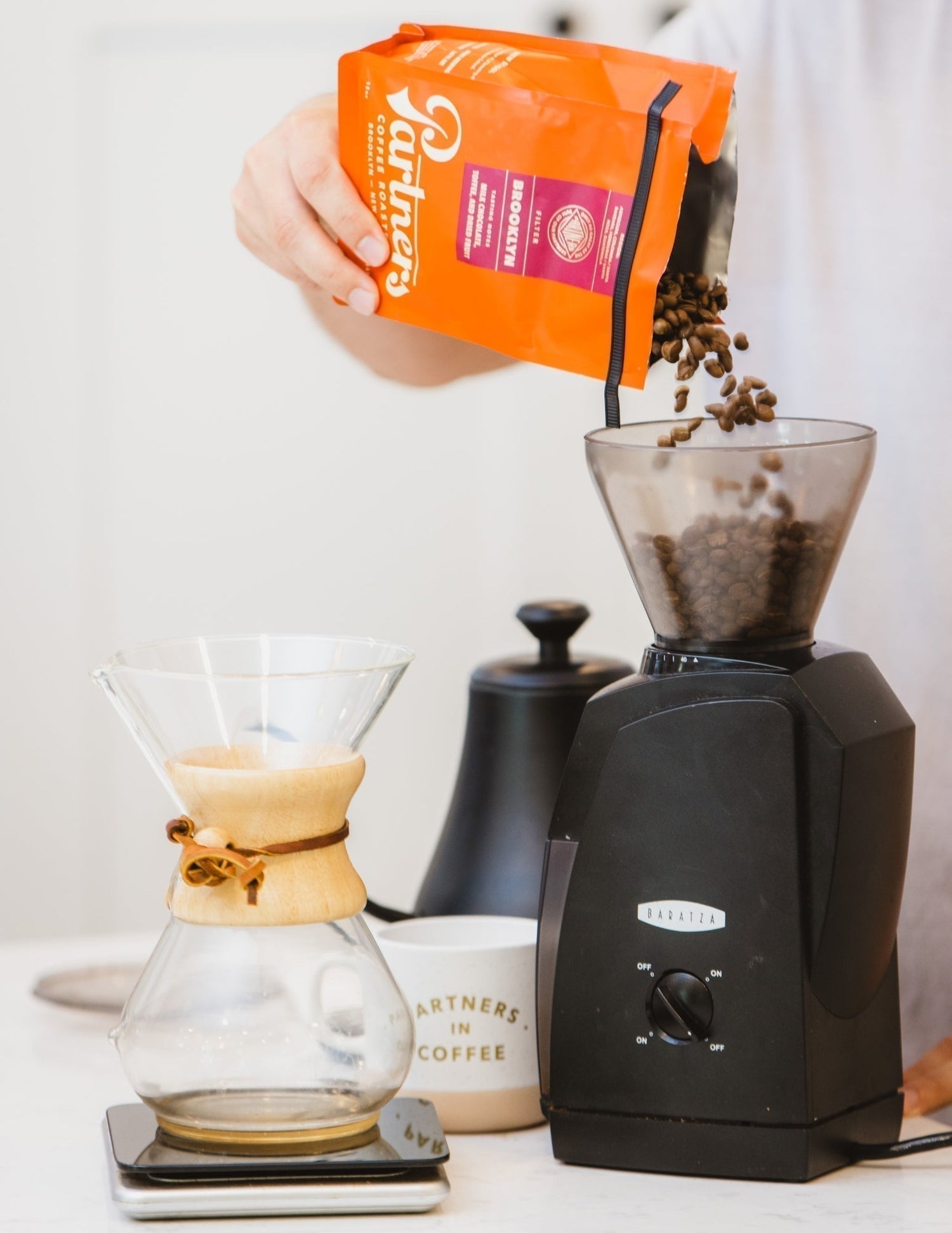 Hand pouring Partners Coffee Brooklyn whole beans into a Baratza grinder beside a Chemex and coffee scale
