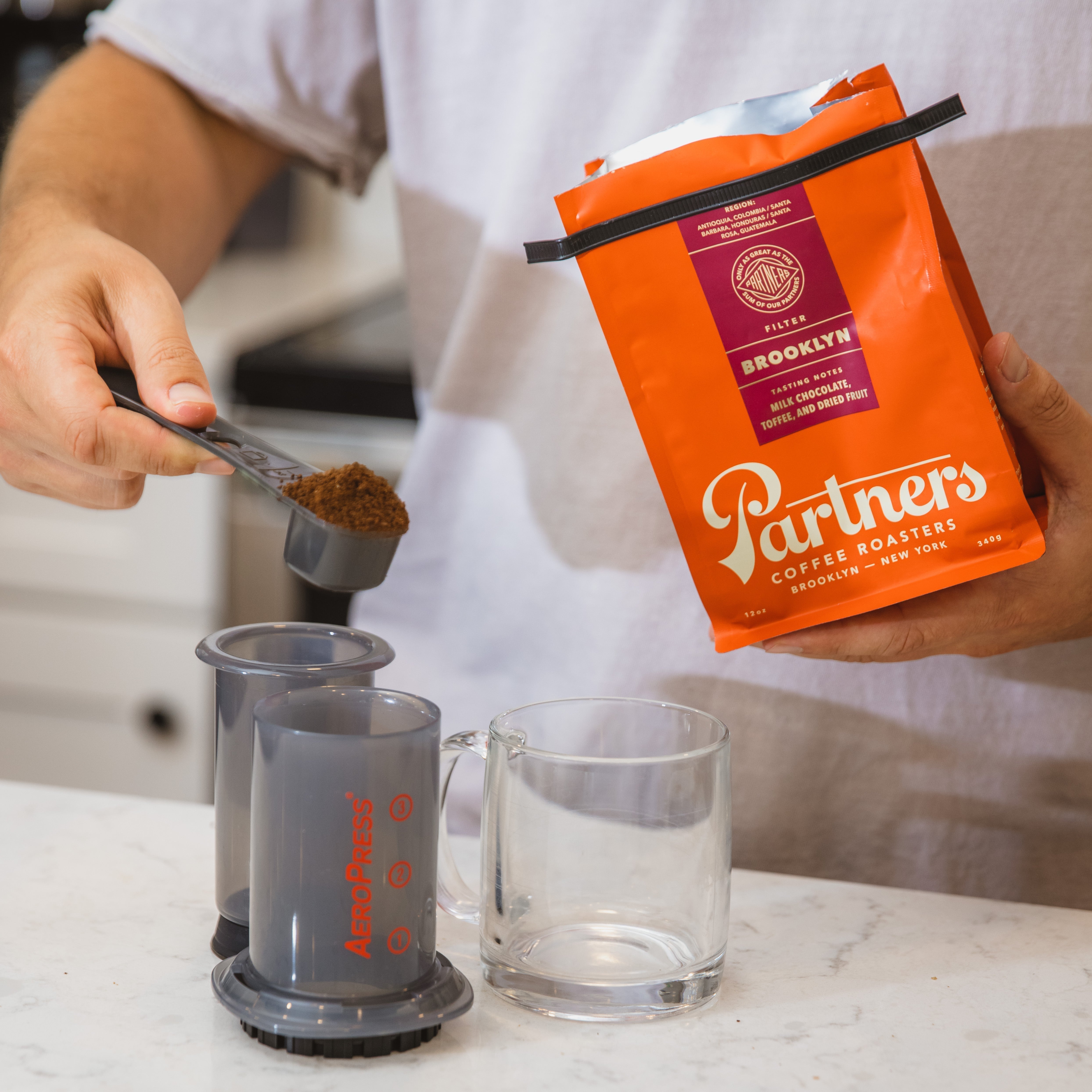 Person scooping Partners Brooklyn ground coffee into an AeroPress with an orange Partners bag held over a kitchen counter