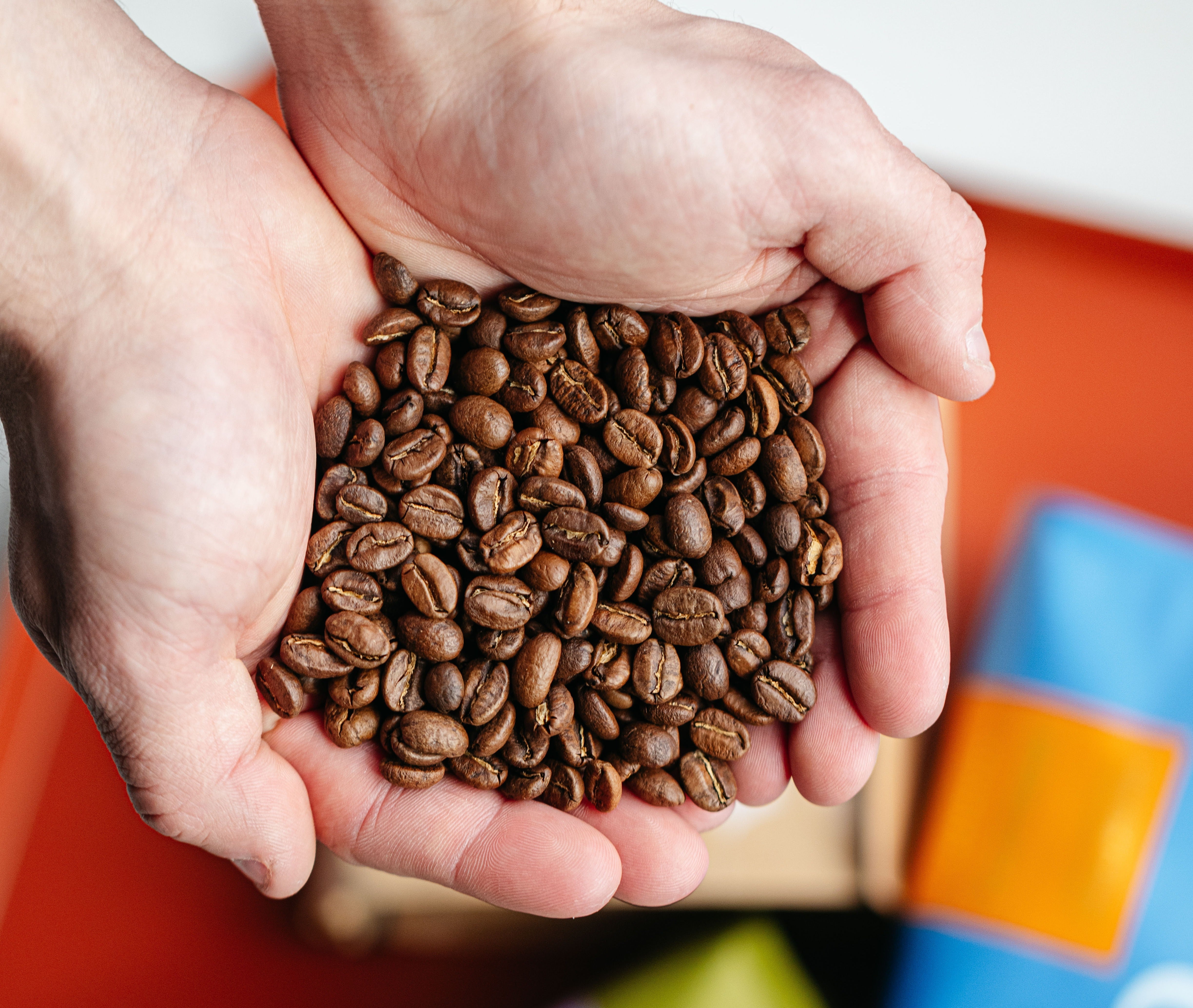 Cupped hands holding freshly roasted coffee beans with blurred colorful packaging in the background