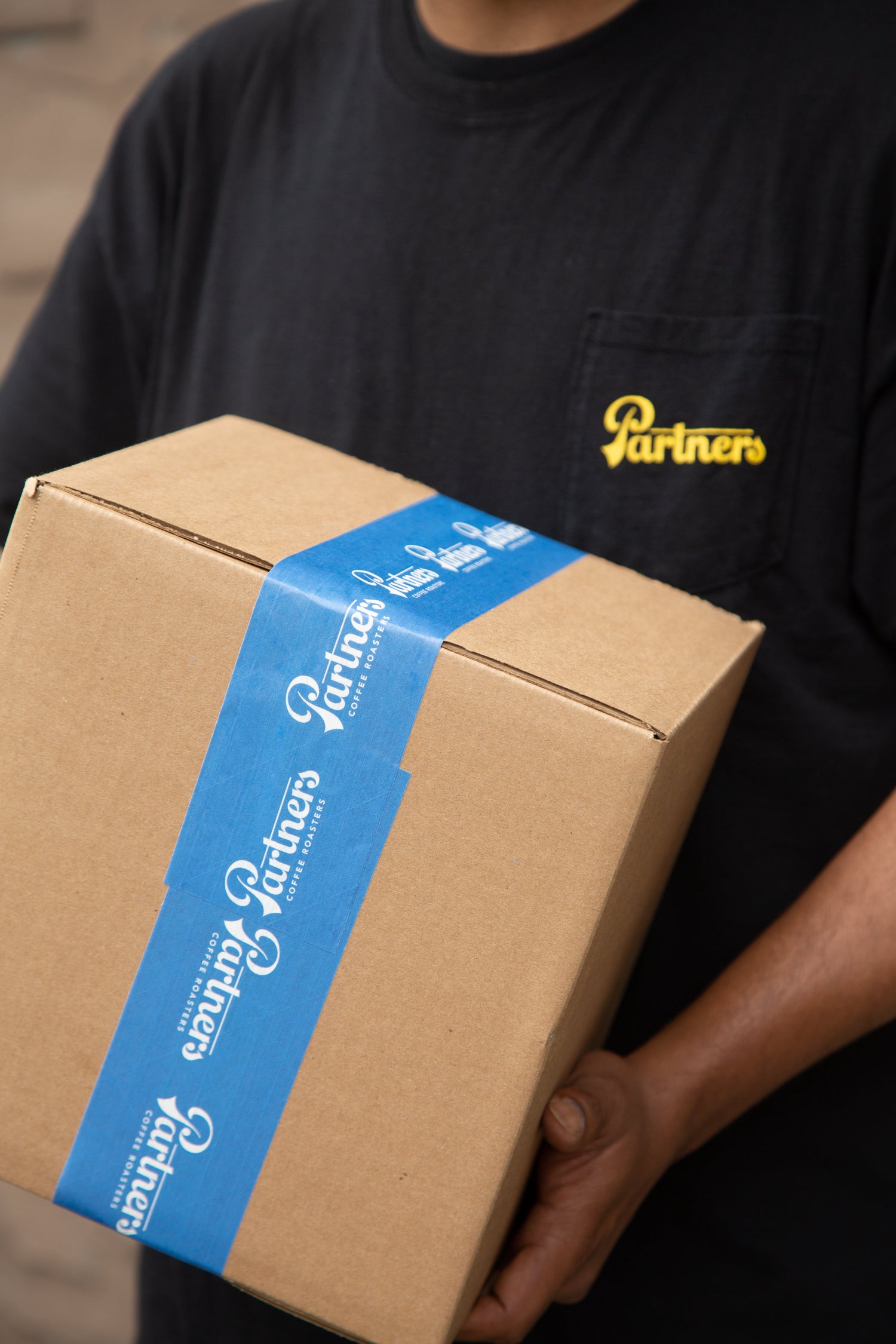 Person holding a cardboard box sealed with blue Partners Coffee Roasters tape, wearing a black shirt with a yellow Partners logo