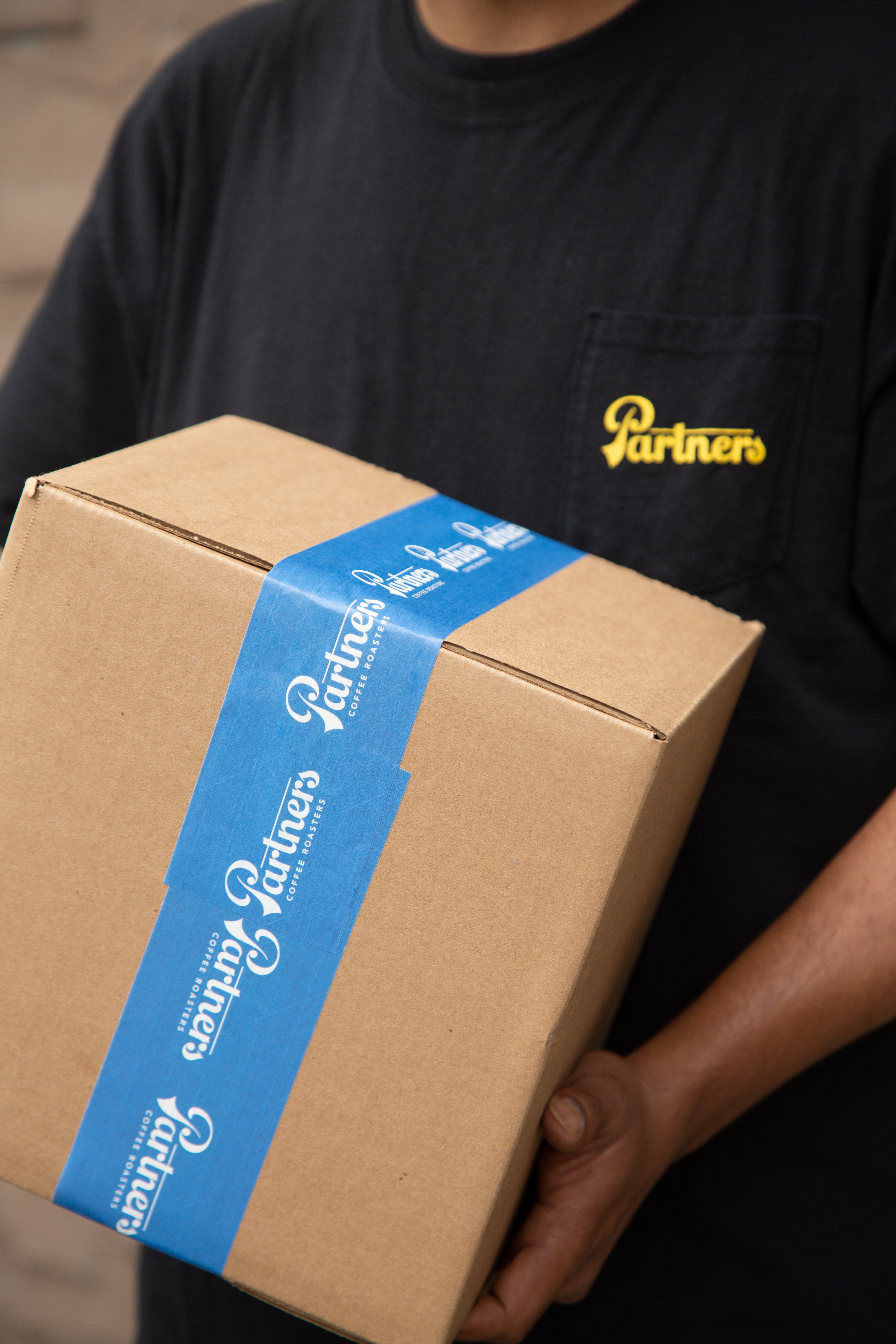 Person holding a cardboard box sealed with blue Partners Coffee Roasters tape, wearing a black shirt with a yellow Partners logo