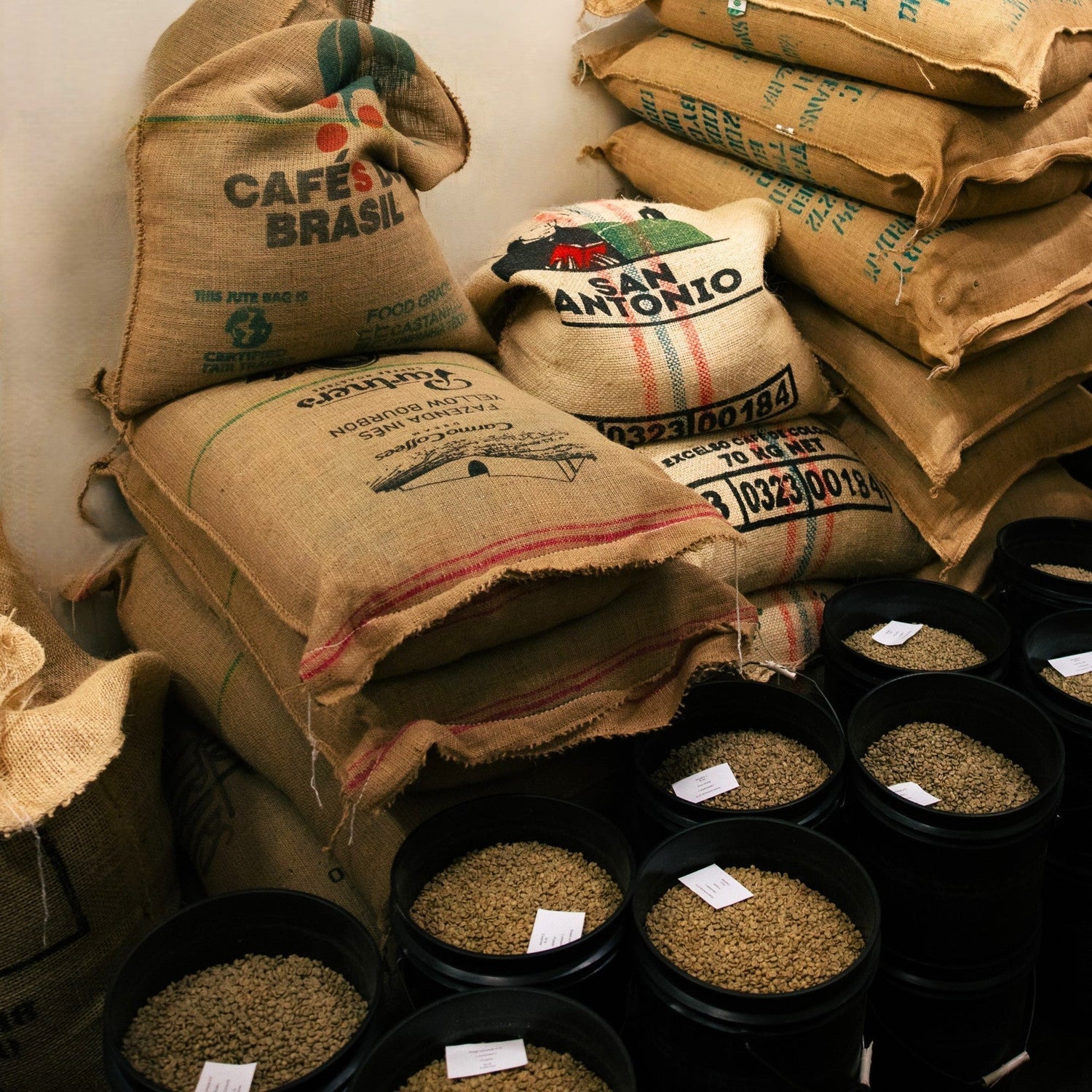 Stacks of burlap coffee sacks and black buckets of unroasted green coffee beans in a roasting storage room