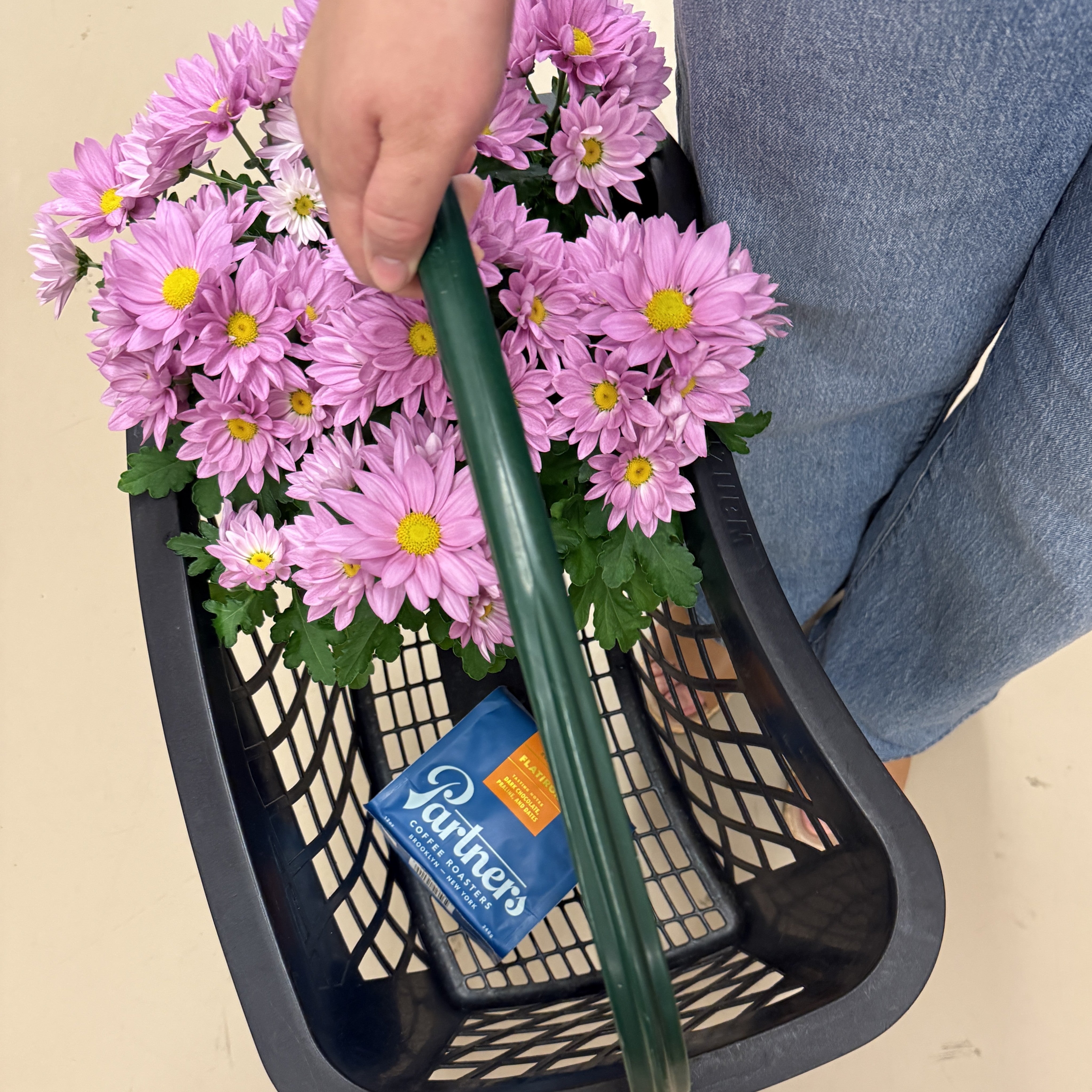 Person holding a shopping basket with pink chrysanthemum flowers and a bag of Partners Coffee Flatiron roast