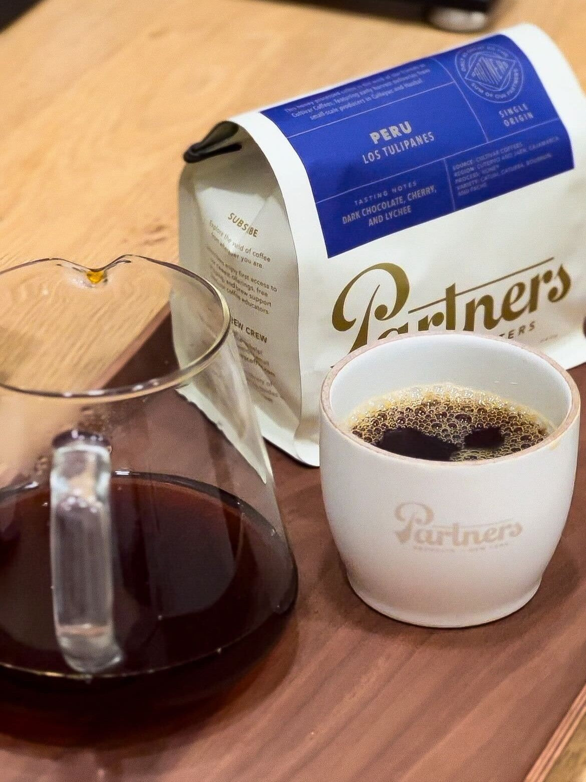 Bag of Partners Coffee Peru Los Tulipanes beside a poured V60 carafe and a Partners mug of brewed coffee on a wooden table