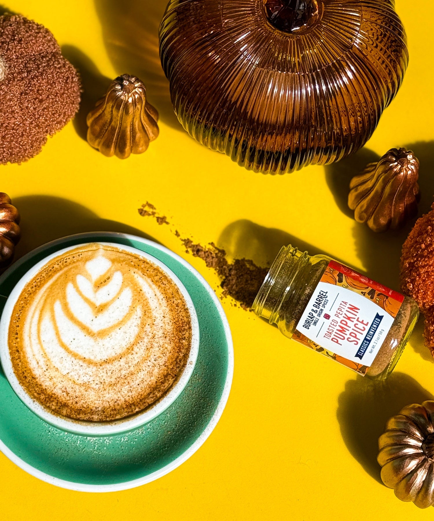 Pumpkin spice latte with leaf latte art beside a spilled jar of pumpkin spice and metallic pumpkins on a yellow background