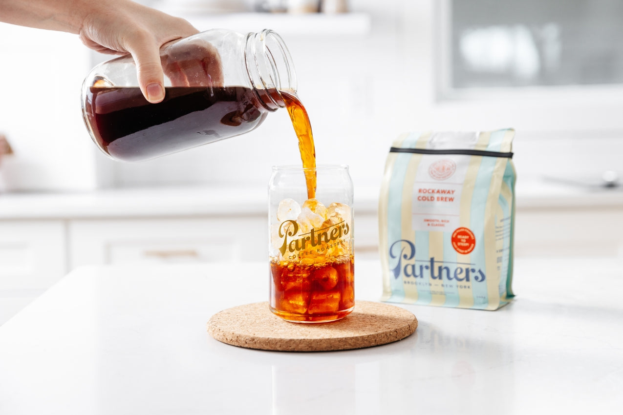 Hand pouring cold brew into an ice-filled Partners glass with a Rockaway Cold Brew pouch on a white kitchen counter