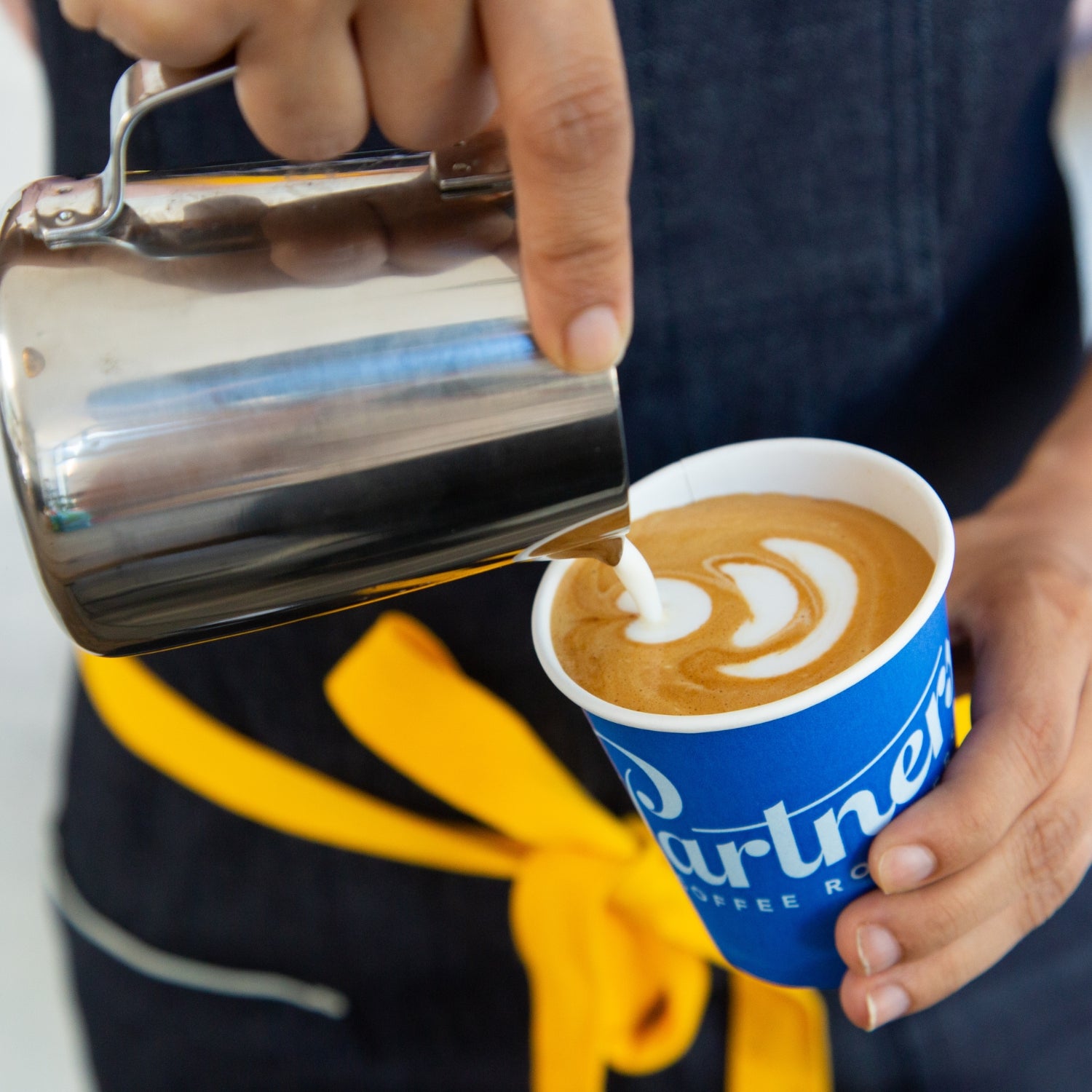 Barista pouring steamed milk to create latte art in a blue Partners Coffee paper cup, close up of hands and stainless steel pitcher