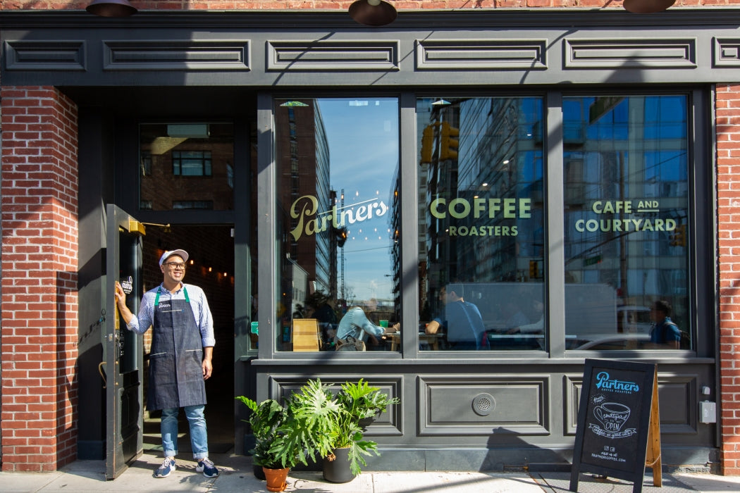 Staff member standing in Partners Coffee doorway; window signs read ‘Partners’, ‘COFFEE ROASTERS’, and ‘CAFE AND COURTYARD’