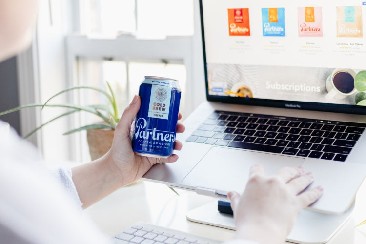 Person holding a Partners cold brew can while browsing the Partners Coffee website on a laptop at a bright desk