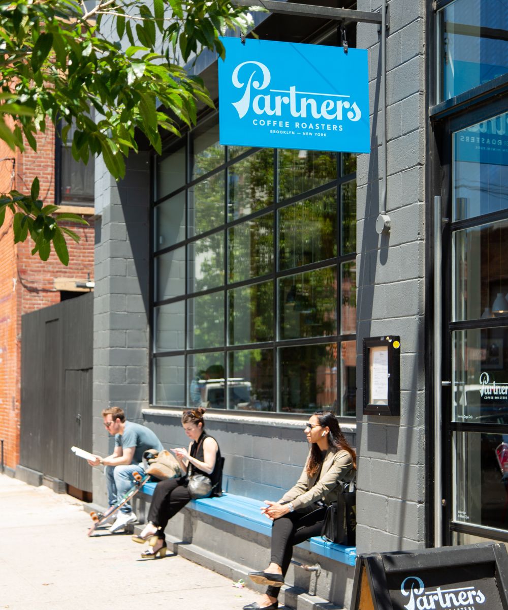 Three people sitting on a bench outside the Partners Coffee Roasters storefront beneath a blue hanging sign