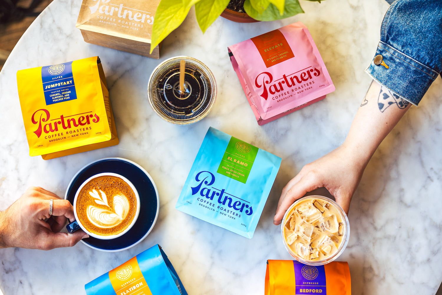 Overhead view of two hands reaching for a latte and an iced coffee surrounded by colorful Partners Coffee bags on a marble table