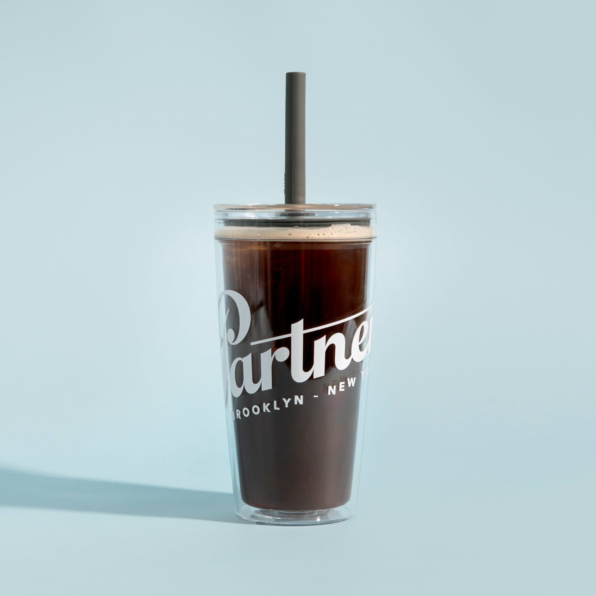 Front of clear MiiR Everywhere straw tumbler with white Partners logo, filled with coffee and gray straw on blue background