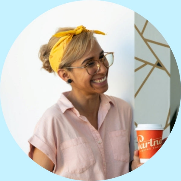 Raina smiling while holding a to-go coffee cup, wearing a yellow headband and glasses against a pale blue circular background