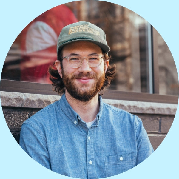 Sam, Partners Coffee green coffee buyer, smiling in front of a brick window wearing a green cap and blue denim shirt