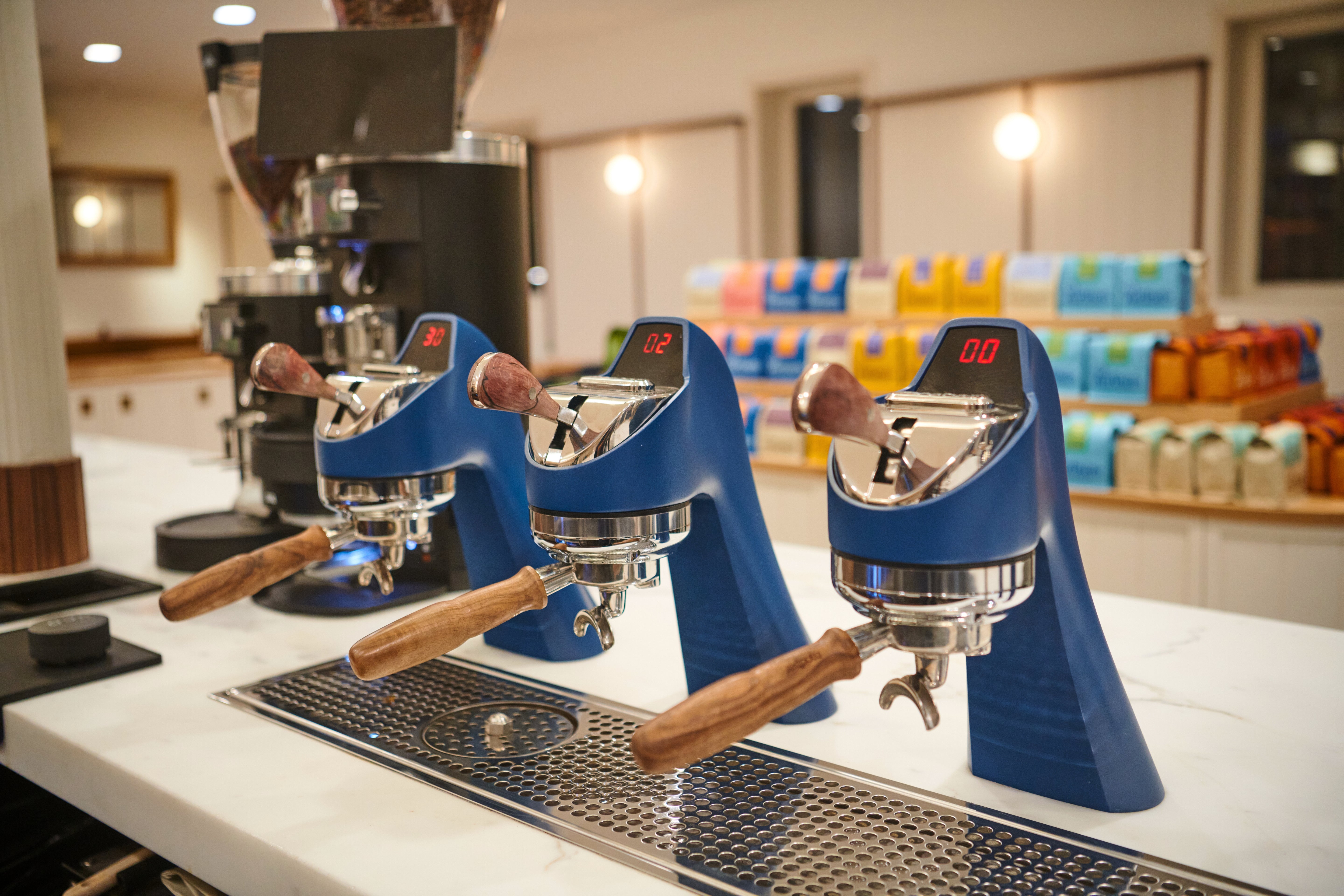 Three blue espresso machines with wooden handles on a white counter at a Partners Coffee cafe, packaged coffee bags on shelves behind