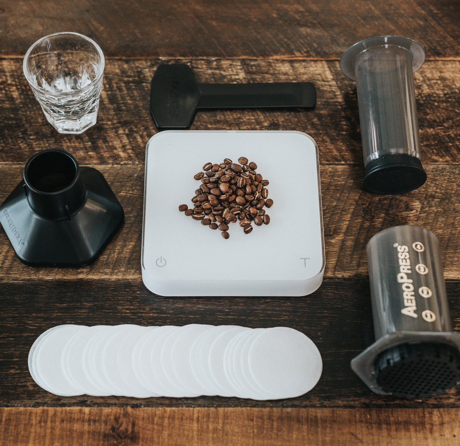 Travel coffee gear laid out on a wooden table: portable brewer parts, digital scale with coffee beans, paper filters, and measuring tools