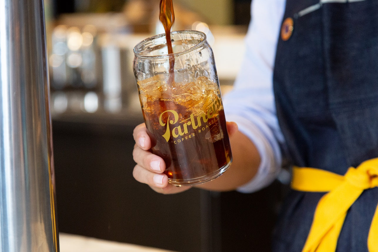 Barista pouring iced coffee into a Partners-branded glass filled with ice at a coffee bar