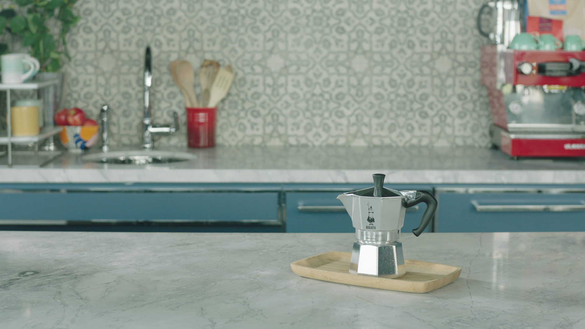 Front view of moka pot showcasing octagonal aluminum body and black handle on a wooden tray on a marble countertop in a kitchen