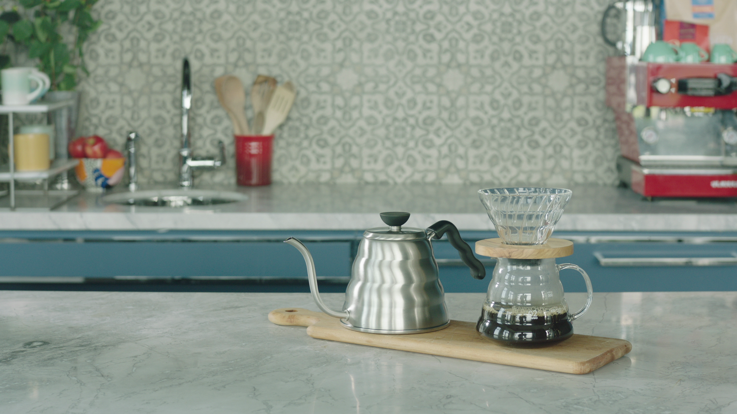 Stainless gooseneck kettle and Hario V60 dripper on a wooden board on a marble kitchen counter with tiled backsplash