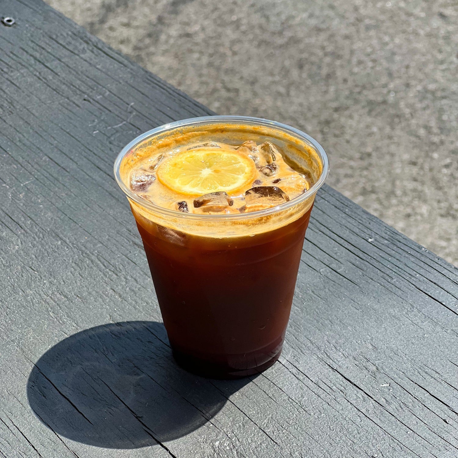 Front view of Citron Espresso Tonic in a clear plastic cup showcasing ice, crema, and a candied orange slice on a sunlit weathered wooden bench