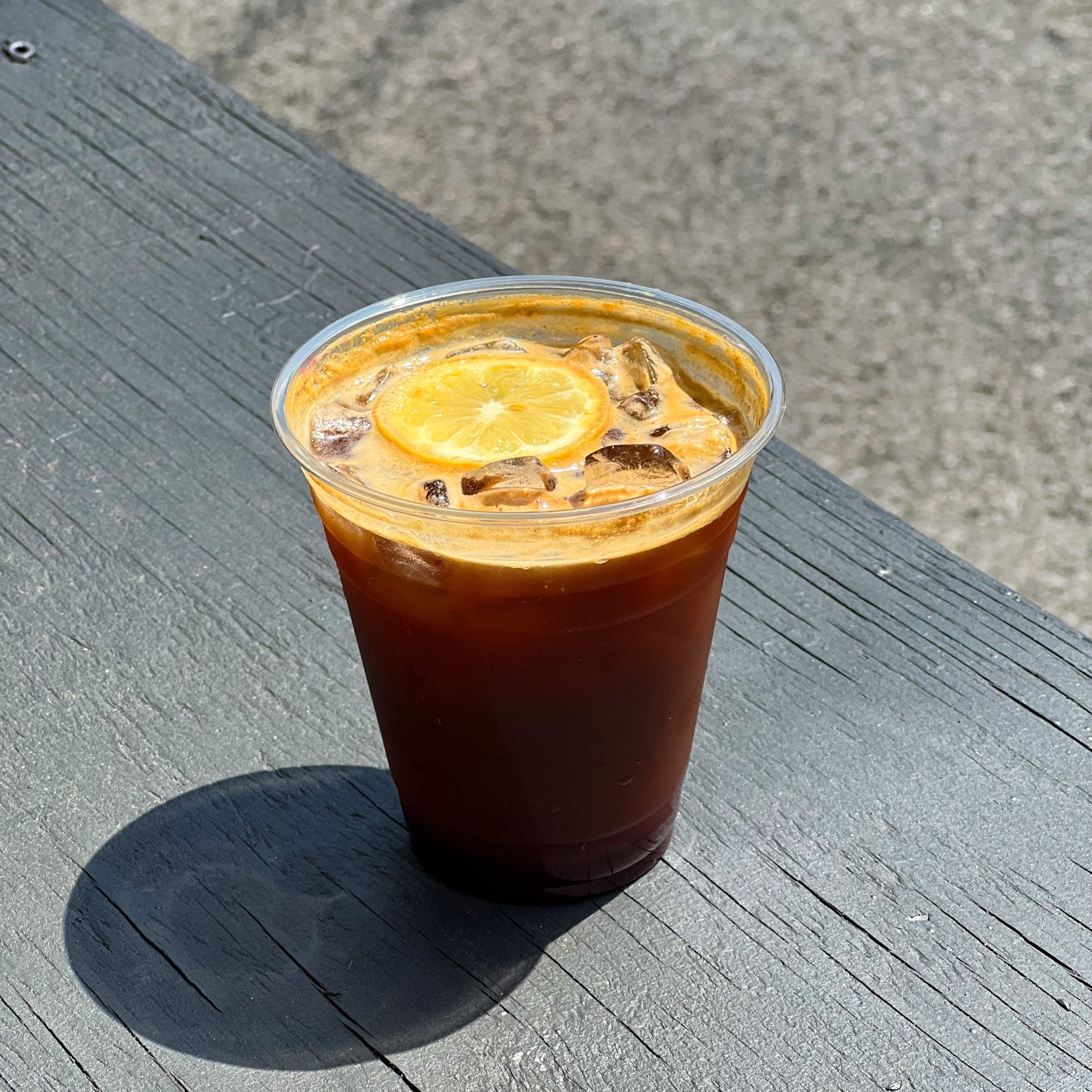 Front view of Citron Espresso Tonic in a clear plastic cup showcasing ice, crema, and a candied orange slice on a sunlit weathered wooden bench