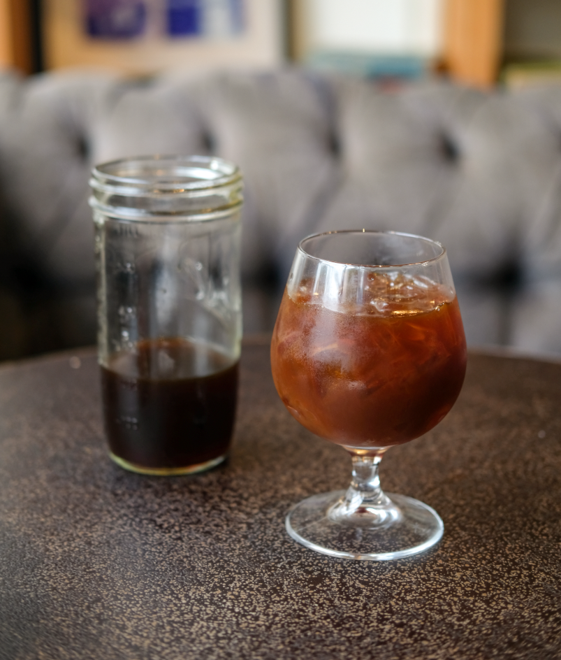 Glass of cold brew whiskey over ice on a small round table, next to a jar of steeped coffee with a tufted sofa in the background