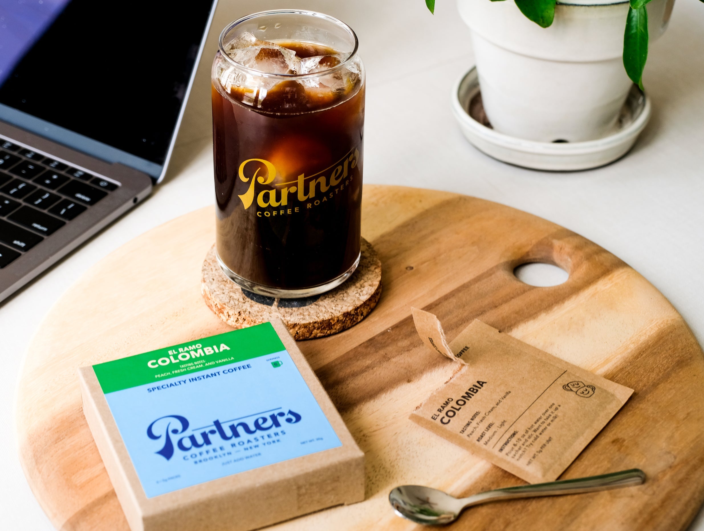 Glass of Partners iced coffee on a cork coaster beside El Ramo Colombia instant coffee sachet and box on a wooden board