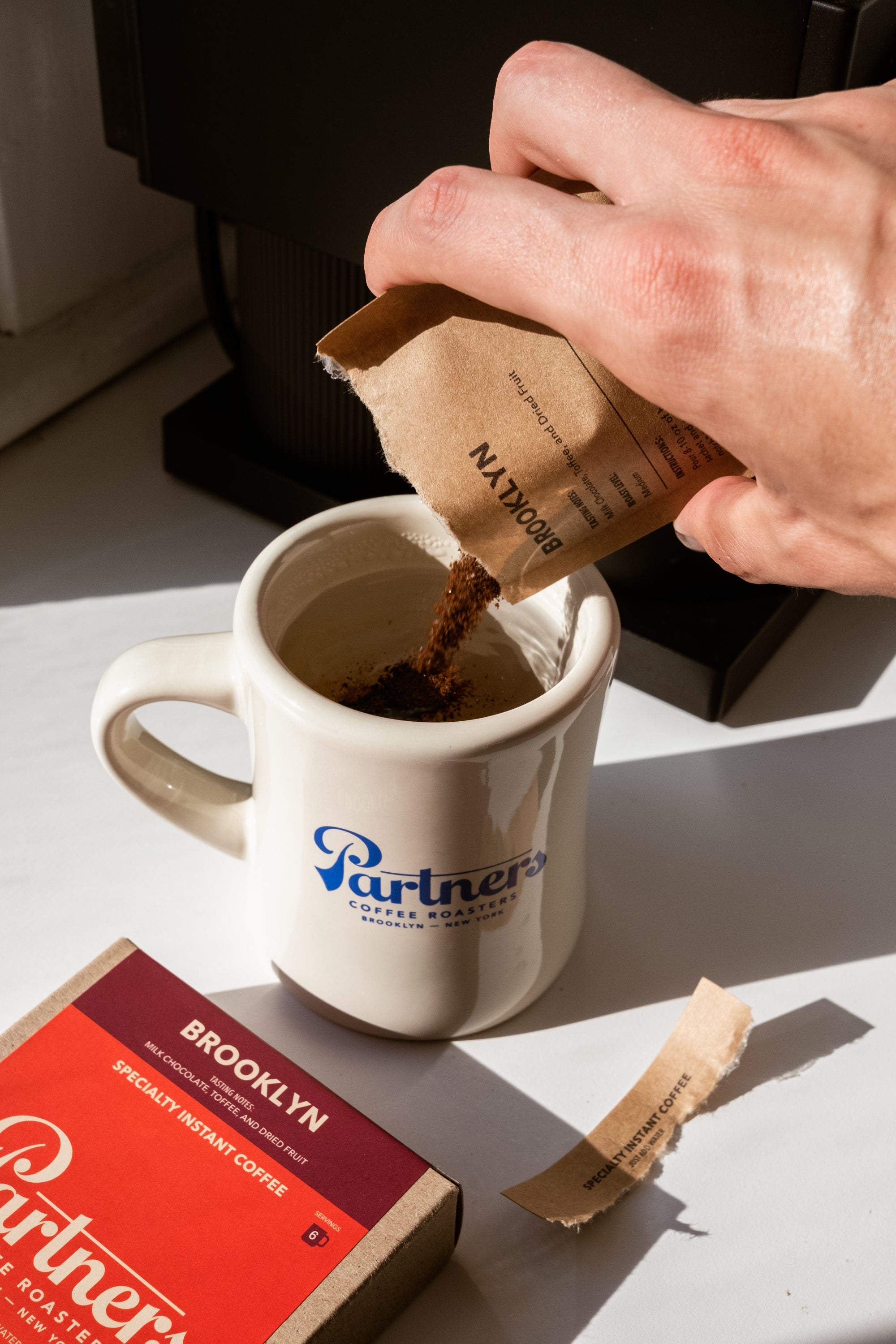 Hand pouring instant coffee into a Partners Coffee mug on a sunlit counter with a Brooklyn instant coffee box nearby