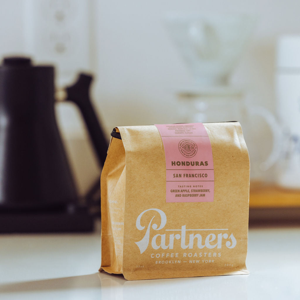 Three-quarter view of Partners Coffee brown bag of Honduras San Francisco natural processed coffee with pink label showing tasting notes, blurred kettle and dripper behind