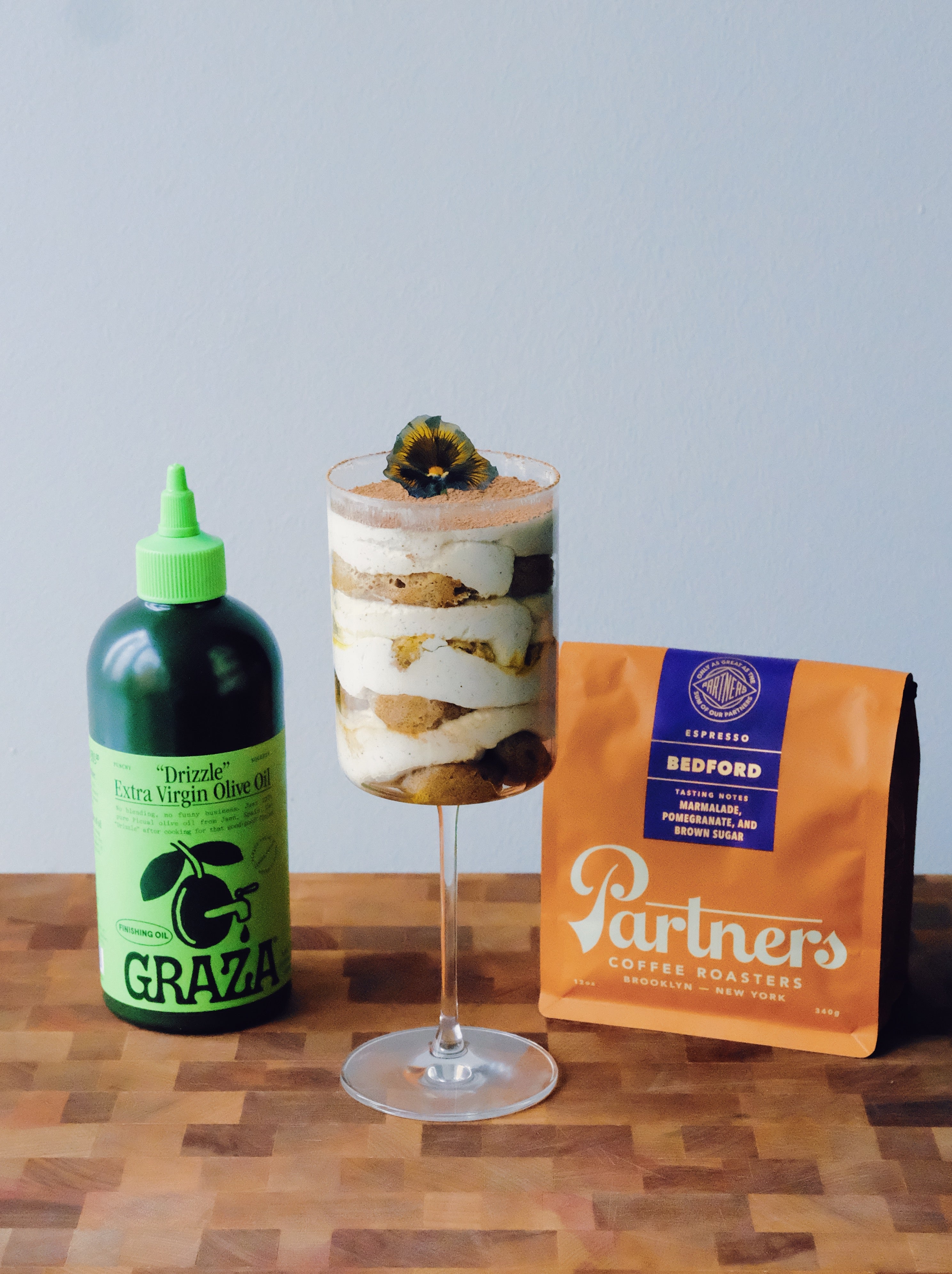 Mini olive oil tiramisu in a stem glass between a green Graza bottle and an orange Partners Coffee Bedford bag