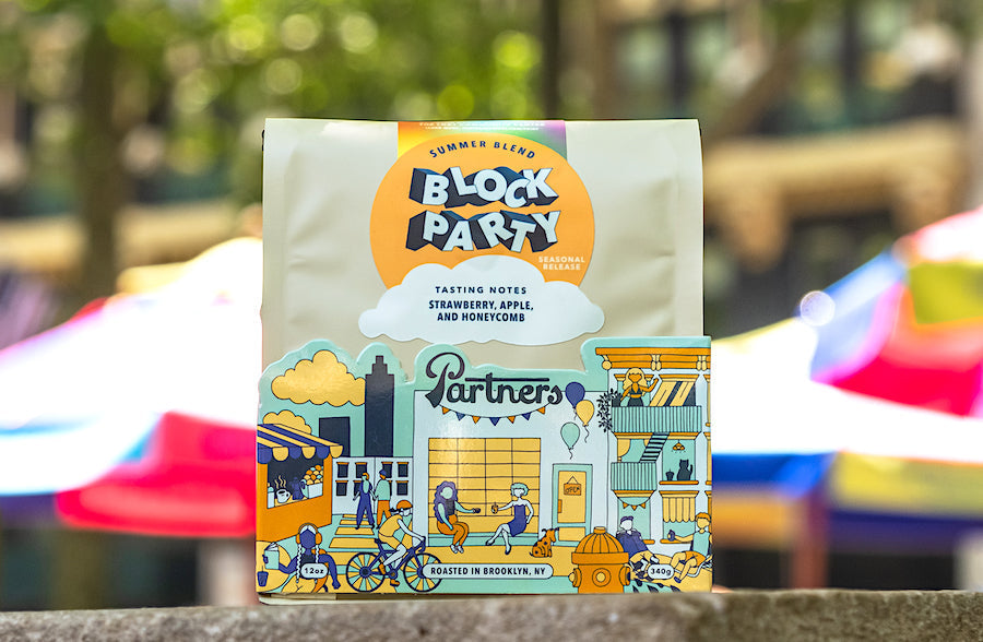 Front view of Block Party summer blend coffee bag showing Block Party logo and tasting notes strawberry, apple, and honeycomb with colorful neighborhood illustration and blurred market umbrellas in the background
