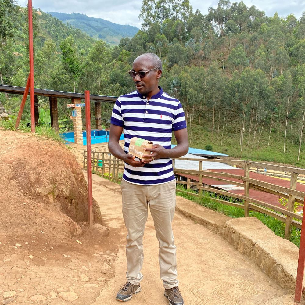 Man holding a bag of Karambi coffee at Karambi Washing Station, standing on a dirt path with drying beds and forested hills behind him