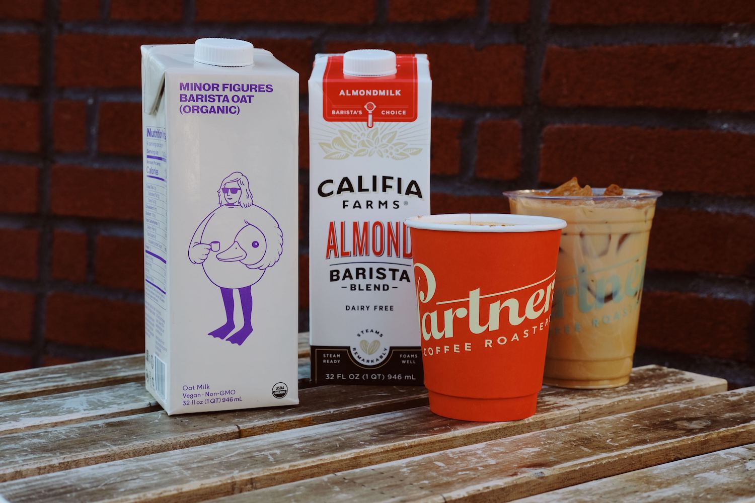Two barista-style plant milk cartons beside a hot Partners Coffee cup and an iced coffee on a weathered wooden bench in front of a brick wall