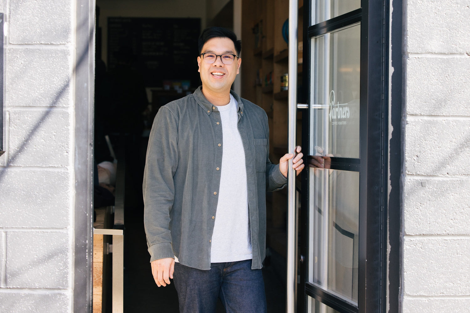 Cary W., Partners Coffee Director of Coffee, smiling as he holds open the cafe’s glass storefront door