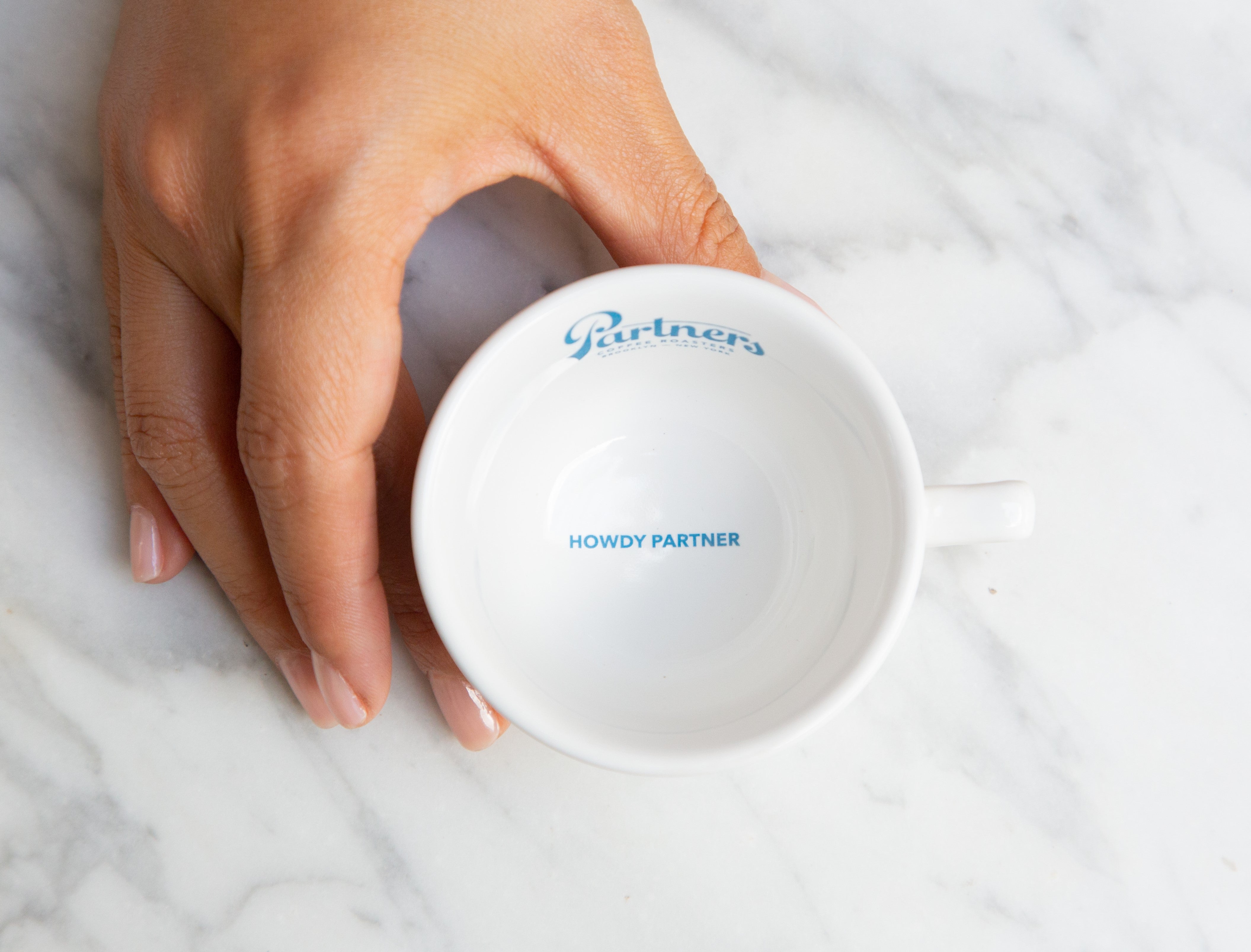 Hand holding a white Partners Coffee espresso cup revealing blue text Howdy Partner inside on a marble countertop