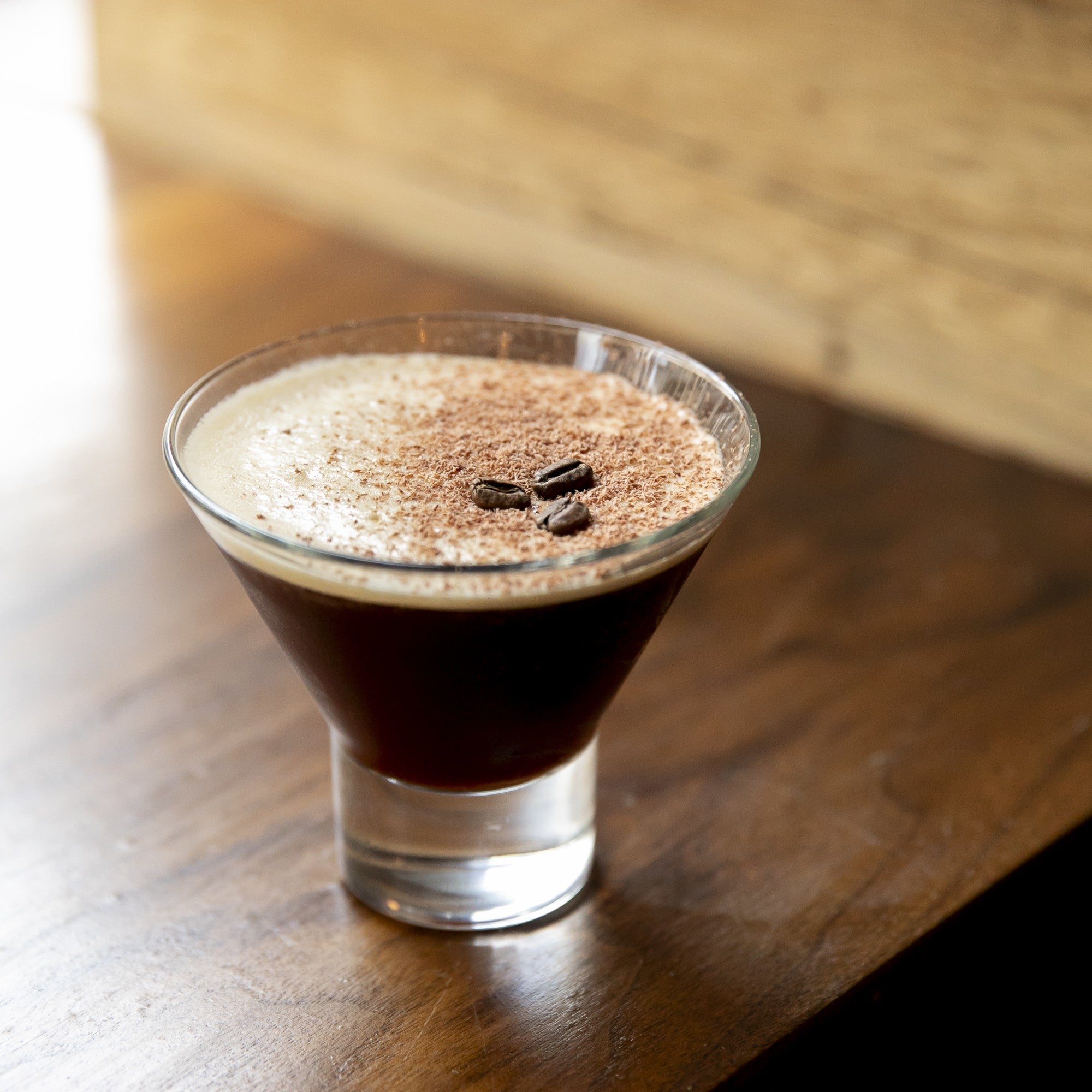 Three-quarter view of an espresso martini showing creamy espresso foam dusted with chocolate and three coffee beans on a wooden table background