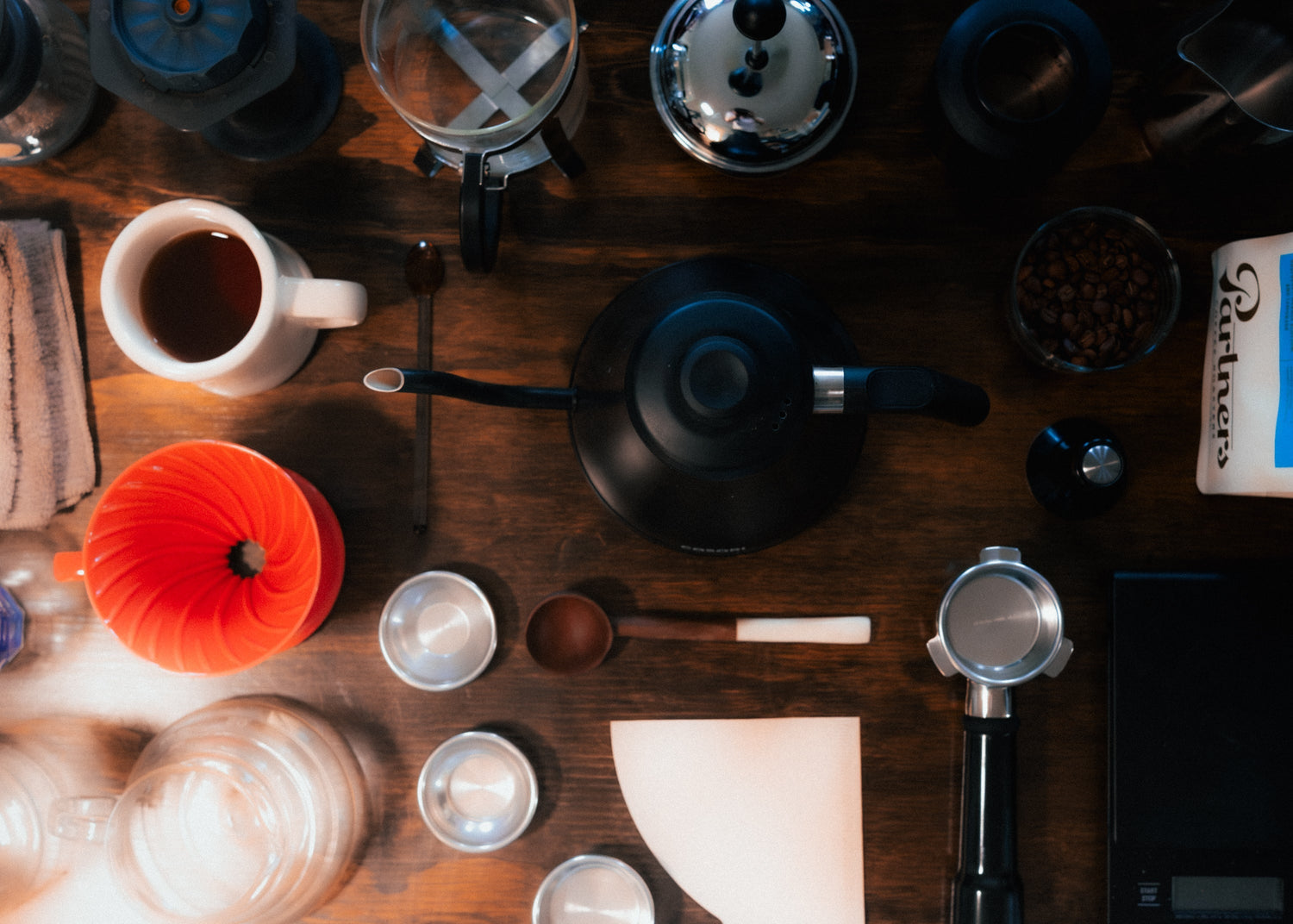 Flatlay of coffee brewing tools on a wooden table with a gooseneck kettle, orange pour-over dripper, portafilter and scale