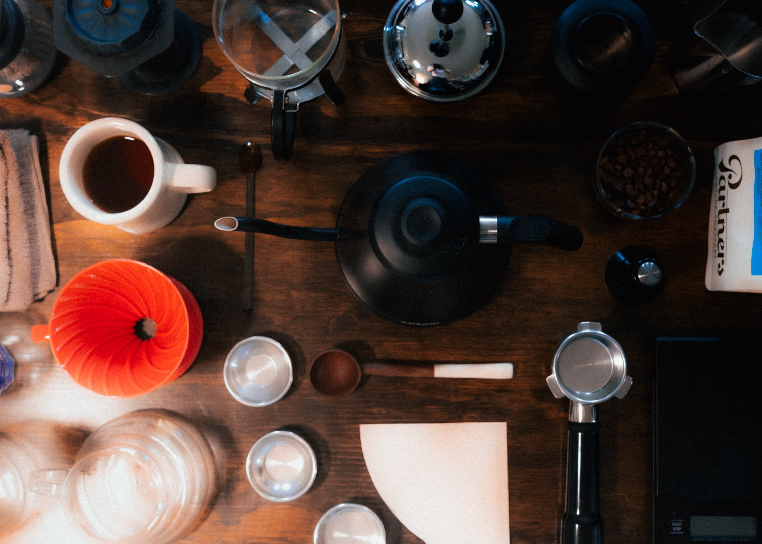 Flatlay of coffee brewing tools on a wooden table with a gooseneck kettle, orange pour-over dripper, portafilter and scale
