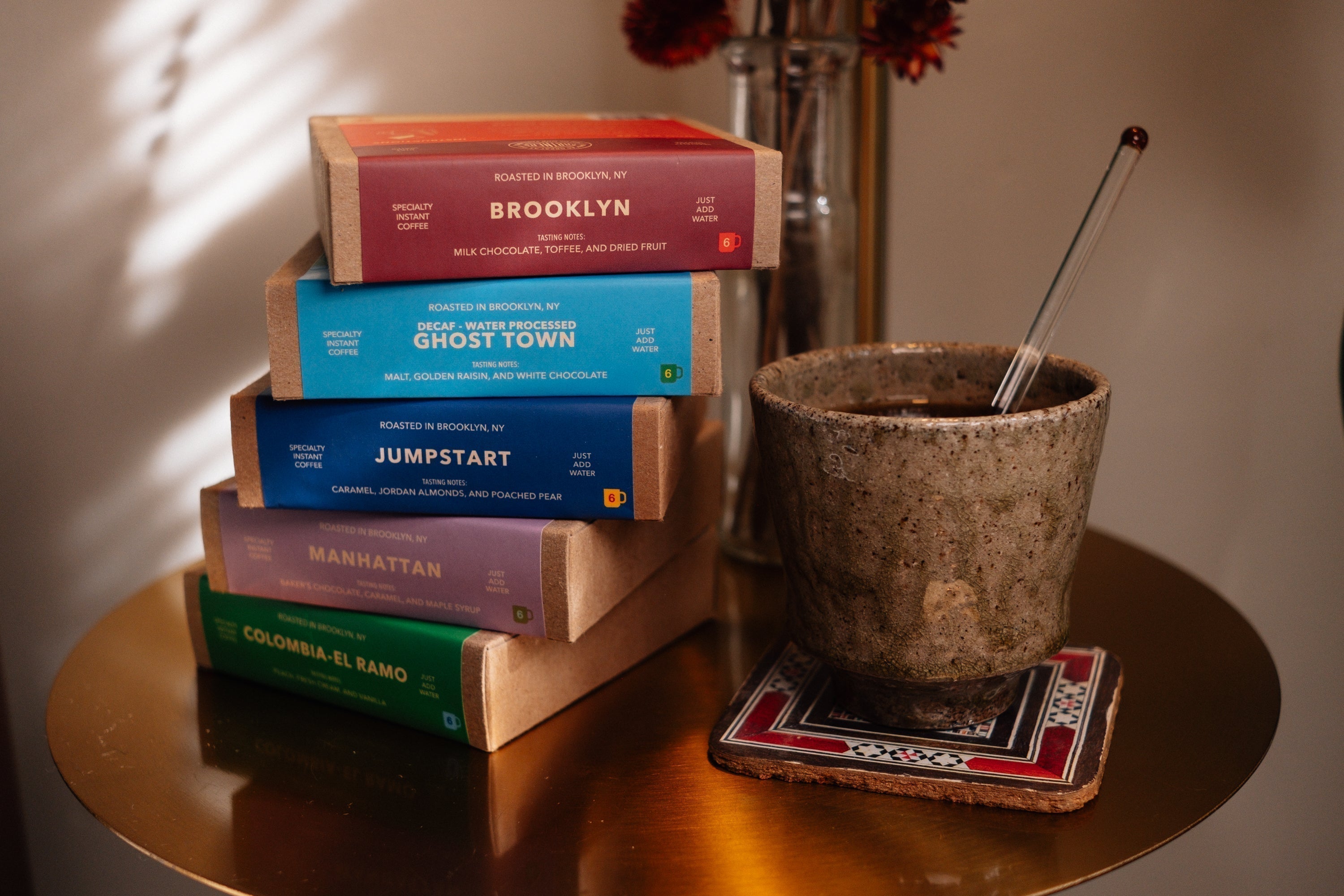 Five stacked Partners Specialty Instant Coffee boxes beside a stone cup of brewed instant coffee on a small round table