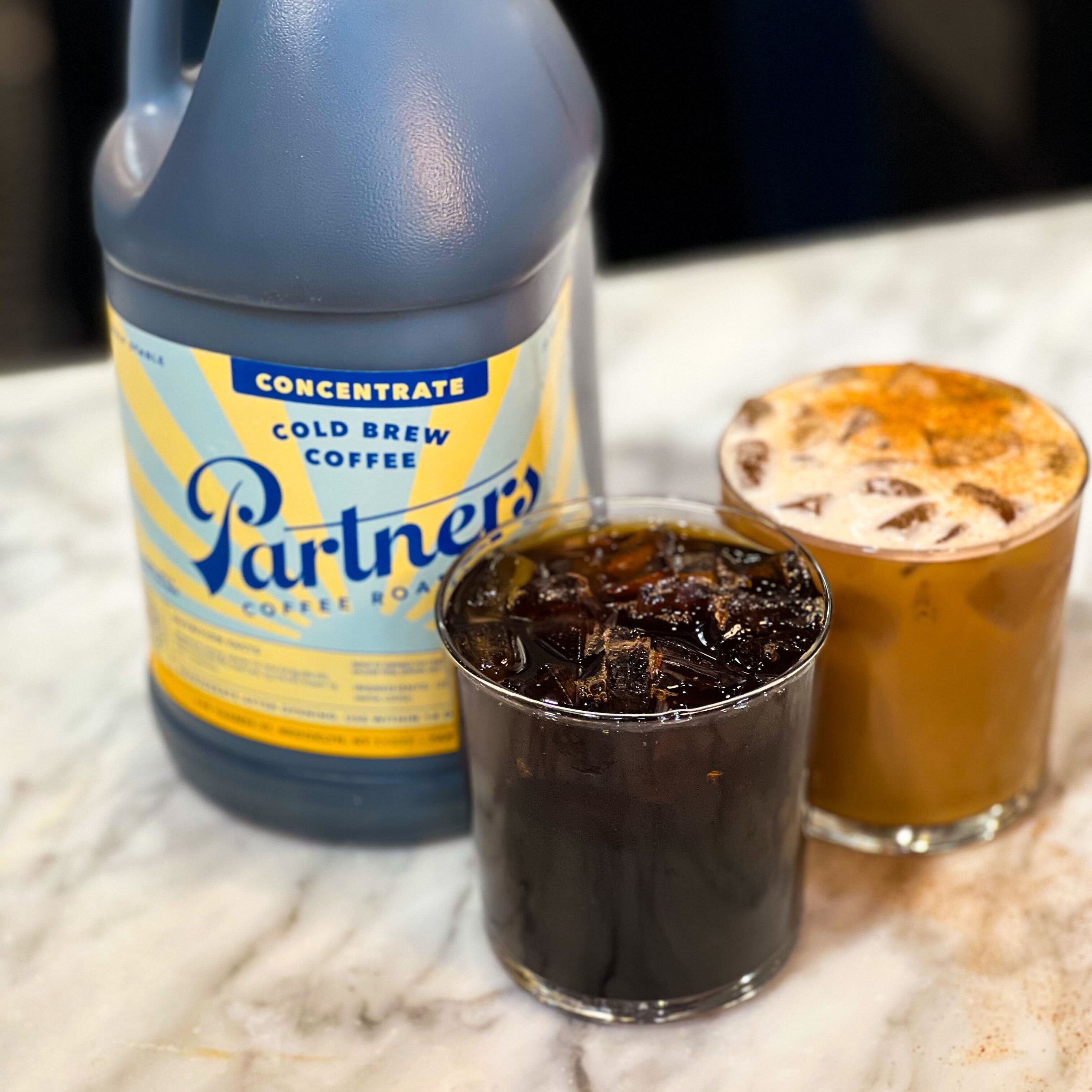 Three quarter view of Partners cold brew concentrate jug with an iced black coffee and an iced latte on a marble countertop