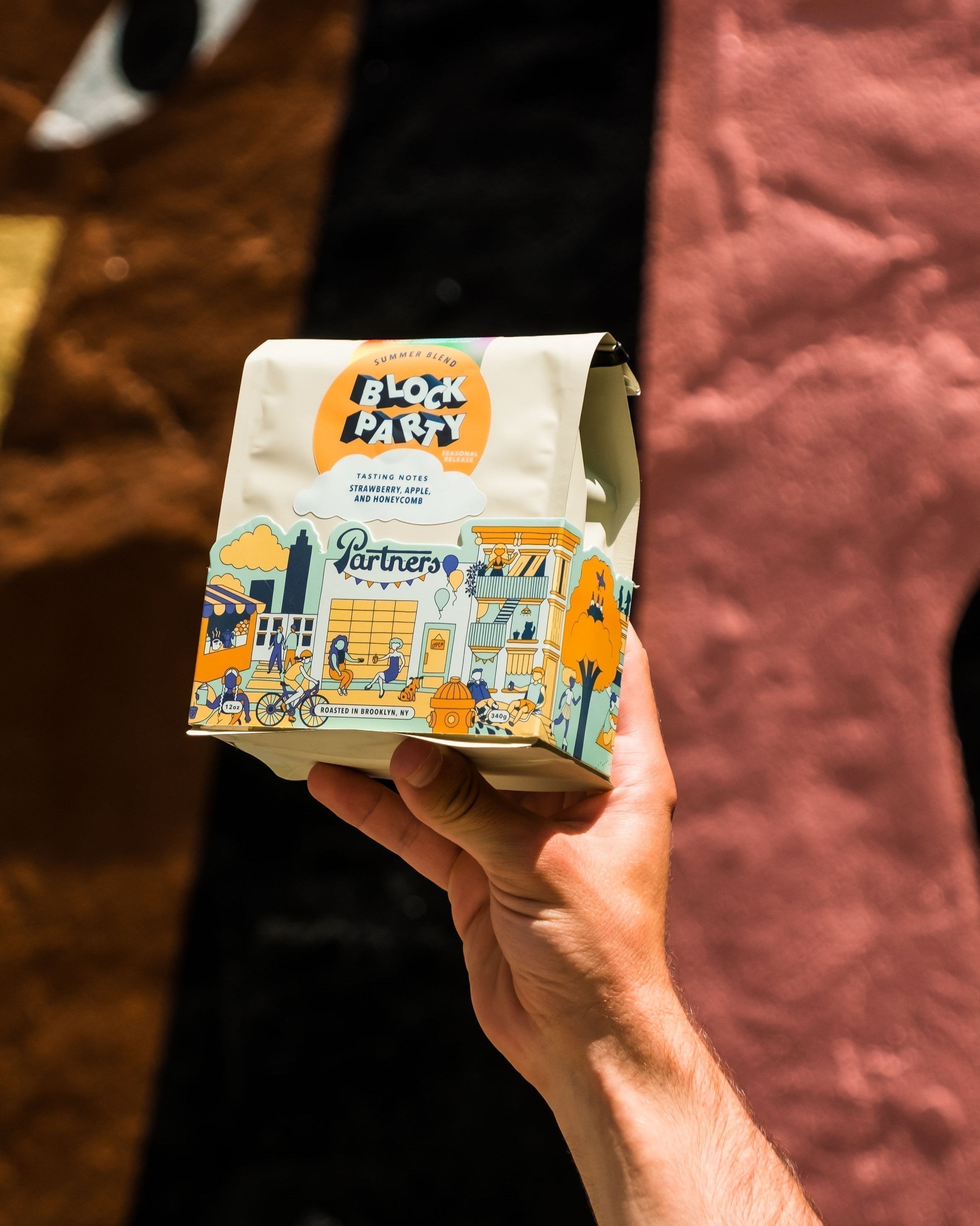 Hand holding Block Party coffee bag with colorful illustrated label and Partners logo against a blurred multicolor backdrop